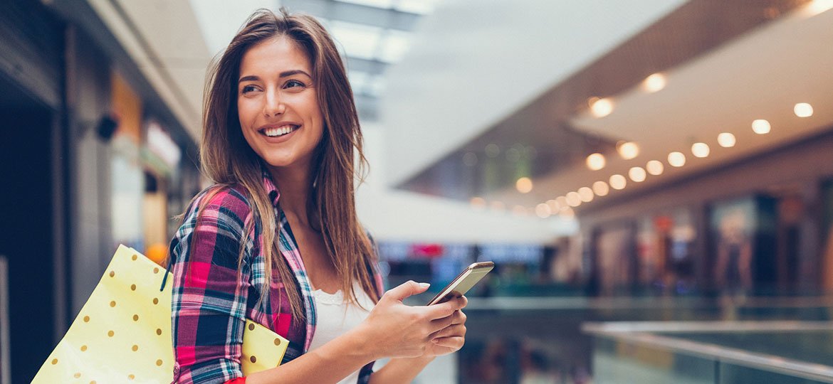 Mulher sorrindo no shopping com sacolas de compra e smartphone