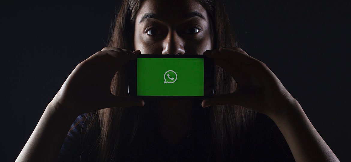 Mulher segurando smartphone em frente a boca, em fundo escuro, com a marca do whatsapp na tela